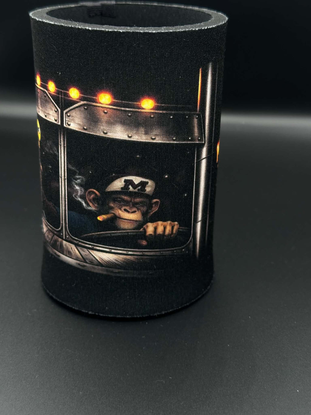 A black neoprene can cooler featuring a graphic print of a chimpanzee wearing a white cap with a black 'M' logo, sitting in a futuristic vehicle cabin with glowing orange lights.